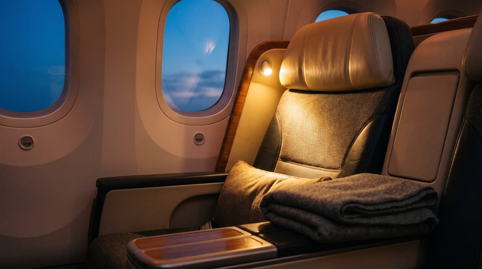 Empty business class window seat with a folded blanket and warm reading light at dusk